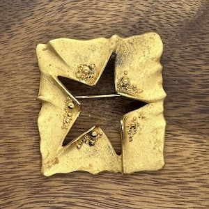 May include: A gold-toned brooch with a star-shaped cutout design. The brooch is on a wooden surface.