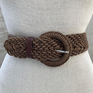 Może przedstawiać: Brązowy pleciony pasek ze sznurka z dużą, okrągłą, srebrną klamrą.