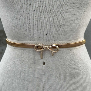 May include: A gold-colored belt with a bow-shaped buckle. The belt is thin and elastic, designed to cinch the waist. The bow buckle adds a decorative element to the belt.