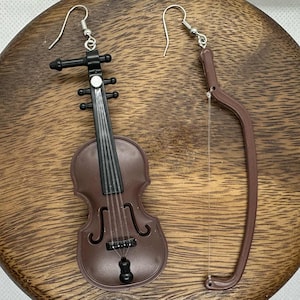 May include: A pair of brown violin-shaped earrings. One earring is a violin, the other is a bow. The earrings have silver-colored hooks. The violin has black strings and a black chin rest. The earrings are on a wooden surface.