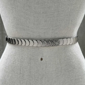 May include: Silver-toned metal belt featuring a series of circular, coin-like embellishments. The belt is worn around the waist of a mannequin, showcasing its design and fit. The belt's design is simple and elegant.