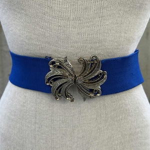 May include: A royal blue elastic belt with a decorative silver-toned buckle. The buckle has an ornate, symmetrical design. The belt is displayed on a mannequin, showcasing its shape and style. This accessory adds a pop of color and vintage flair.