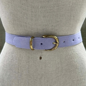 May include: A light purple belt with a gold-colored buckle is displayed on a mannequin. The belt is made of a textured fabric and has a classic design. The gold buckle adds a touch of elegance to the belt.