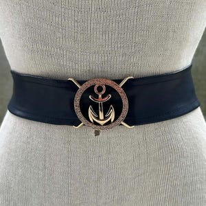 Może przedstawiać: Czarny pasek ze złotą klamrą w kształcie kotwicy. Szeroki pasek jest prezentowany na manekinie. Klamra kotwicy ma okrągłą ramę z kotwicą pośrodku. Dodatek o tematyce żeglarskiej.