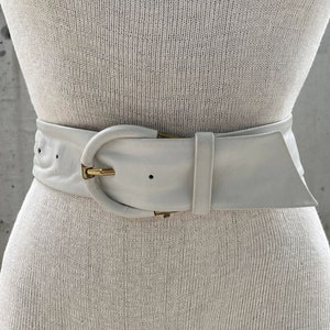 Puede incluir: Un cinturón de cuero blanco con una hebilla dorada. El cinturón es ancho y tiene una forma ligeramente curvada.