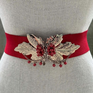 May include: A red elastic belt with a gold-colored leaf and berry clasp. The clasp features detailed leaf designs and red berry clusters, with small red beads hanging from the bottom. The belt is displayed on a mannequin.