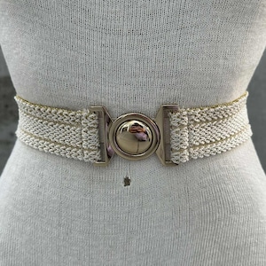 May include: A cream-colored woven belt with a gold-tone buckle. The belt is displayed on a mannequin. The buckle is a large, round, gold-tone metal piece with a clasp closure. The woven material has a subtle gold thread.