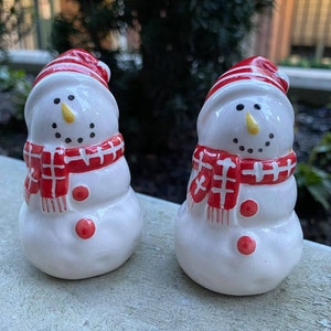 Puede incluir: Dos saleros y pimenteros de cerámica blanca en forma de muñeco de nieve con sombreros y bufandas a rayas rojas y blancas. Los muñecos de nieve tienen ojos negros, una nariz de zanahoria amarilla y botones rojos.