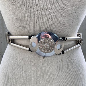 Może przedstawiać: Srebrny pasek z dużą, okrągłą, ozdobną klamrą. Klamra ma wypukły wzór i błyszczące wykończenie.