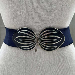 Puede incluir: Un cinturón elástico azul marino con una hebilla geométrica plateada y negra. La hebilla tiene forma de lazo.