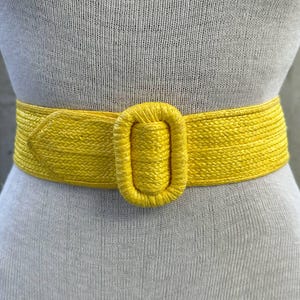 Może przedstawiać: Jasny żółty, tkany pasek z dużą, prostokątną klamrą. Pasek wykonany jest z grubego, fakturowanego materiału i idealnie nadaje się do dodania odrobiny koloru do każdego stroju.