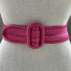 May include: A vibrant fuchsia belt with a woven texture and a matching oval buckle. The belt is wrapped around a mannequin, showcasing its design and fit. This accessory adds a pop of color and style to any outfit.
