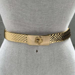 Może przedstawiać: Złoty łańcuszkowy pasek z klamrą w kształcie kwiatu. Pasek wykonany jest z połączonych ze sobą metalowych ogniw.