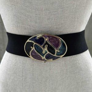 Może przedstawiać: Czarny elastyczny pasek z dużą owalną klamrą. Klamra ma złotą ramę i ma kolorowy wzór z fioletowymi, niebieskimi i turkusowymi kształtami. Pasek jest prezentowany na manekinie.