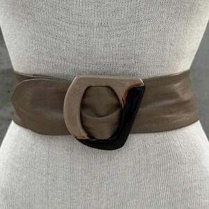 May include: A wide, taupe-colored belt with a large, geometric buckle. The buckle features a combination of tan, black, and gold tones. The belt is shown on a mannequin.