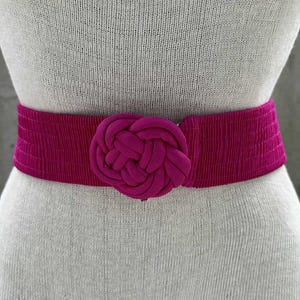 Może przedstawiać: Szeroki, magentowy pasek z dużym, wiązanym detalem z tkaniny pośrodku. Pasek wykonany jest z prążkowanego materiału i prezentowany jest na manekinie. Ten modny dodatek dodaje koloru. Pasek ma około 80-90 cm długości.