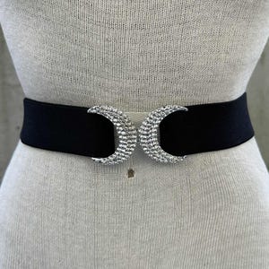 Może przedstawiać: Czarny elastyczny pasek ze srebrną klamrą w kształcie półksiężyca. Klamra jest ozdobiona małymi, błyszczącymi dżetami. Pasek jest prezentowany na manekinie.