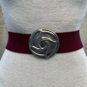 May include: A wide burgundy belt with a large, silver-toned, circular buckle. The buckle features a swirling, three-part design. The belt is displayed on a mannequin, showcasing its shape and style. This accessory is a fashion statement.