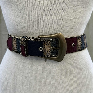 May include: A patterned belt with a large, antique brass buckle. The belt features a design with burgundy, navy, and tan colors. The belt is adorned with small, silver-colored studs along the edges.