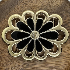 May include: A gold-toned metal belt buckle with a black floral design. The buckle has a flower-like shape with black petals and a gold center. The buckle is set against a wooden background.