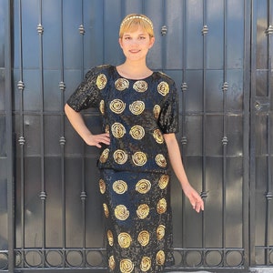 May include: Black sequined two-piece outfit with gold spiral designs. The top has short sleeves and a round neckline. The skirt is knee-length. The outfit is paired with gold-colored heels. A pearl headband completes the look. The outfit is suitable for a party or special event.