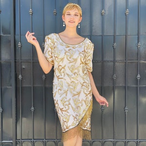 May include: A white, knee-length dress with gold embellishments and short sleeves. The dress features a gold fringe at the hemline and is adorned with intricate gold patterns. The dress is paired with neutral-colored heels.