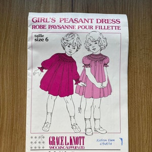 Puede incluir: Un patrón de costura para un vestido campesino para niña, talla 6, con un corpiño de smock rosa y un cuello blanco. El patrón es de Grace L. Knott Smocking Supplies Ltd. e incluye instrucciones para el smocking inglés.