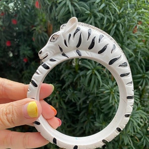 Puede incluir: Brazalete de brazalete blanco y negro con temática de cebra. El brazalete presenta un diseño de cabeza de cebra con rayas negras y una forma curva. El brazalete está hecho de un material sólido y tiene aproximadamente 7.6 cm de diámetro.