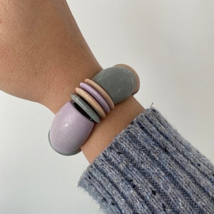Puede incluir: Un brazalete hecho de varias cuentas grandes, redondas y coloridas. Las cuentas son en tonos de morado, rosa y gris.