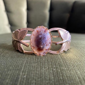 Może przedstawiać: Złota bransoletka z dużym, owalnym, brązowo-białym agatem osadzonym w środku. Bransoletka ma dwa ozdobne, liściaste akcenty po obu stronach kamienia.