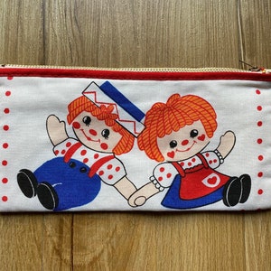 Puede incluir: Un estuche para lápices de lona blanca con lunares rojos y cierre de cremallera. El estuche presenta dos muñecas de pelo rojo con vestidos de lunares rojos y blancos y pantalones azules. Una muñeca lleva un gorro de marinero azul y blanco.