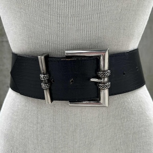May include: A wide black belt with a textured surface and a large silver-colored buckle. The buckle is square and has decorative rope-like accents. The belt is displayed on a mannequin.