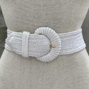 Może przedstawiać: Biały tkany pasek z dużą, okrągłą klamrą. Pasek wykonany jest z teksturowanego materiału i posiada złotą metalową klamrę. Pasek jest pokazany na manekinie.