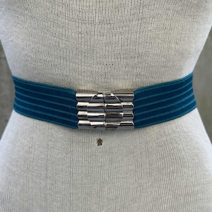 May include: Teal velvet belt with a silver-tone buckle. The belt is worn on a mannequin. The buckle is rectangular with a series of horizontal bars. The velvet belt is approximately 1.5 inches wide.