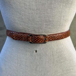 May include: A brown snakeskin belt with a rectangular silver buckle is displayed on a mannequin. The belt is thin and has a textured pattern. The mannequin is a neutral colour, and the background is blurred.