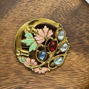 Puede incluir: Un broche dorado con diseño floral. El broche presenta flores de esmalte rosa, verde y azul con diamantes de imitación transparentes y azules.