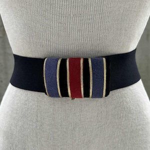May include: A black elastic belt with a decorative center. The center features vertical stripes of blue, red, and gold. The belt is shown on a mannequin.