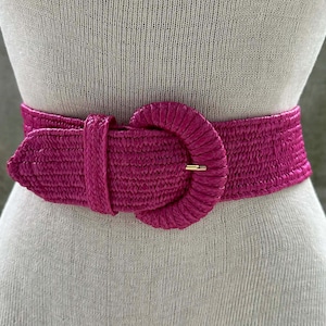 May include: A vibrant fuchsia belt with a wide woven design. The belt features a large, circular buckle with a gold-colored metal clasp. The belt is displayed on a mannequin, showcasing its shape and style.
