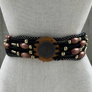 Może przedstawiać: Szeroki pas z drewnianą klamrą i ozdobnymi koralikami. Pas posiada czarny, tkany pas z złotymi akcentami i rzędami brązowych, owalnych koralików. Drewniana klamra ma okrągły wzór ze szczegółowym wzorem.