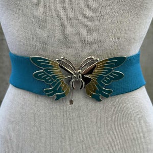 Puede incluir: Cinturón elástico turquesa con cierre de mariposa plateado. La mariposa tiene alas turquesas y doradas. El cinturón se muestra en un maniquí. Este cinturón es un accesorio de moda.