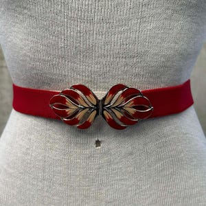 Może przedstawiać: Czerwony elastyczny pasek z ozdobnym metalowym zapięciem. Zapięcie ma wzór liści z czerwonymi, kremowymi i srebrnymi akcentami. Pasek ma szerokość około 3,8 cm.