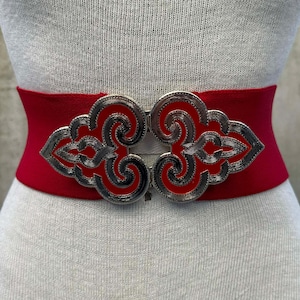May include: A red elastic waist belt with a decorative silver-colored clasp. The clasp features a cloud-like design with red and black accents. The belt is displayed on a mannequin.