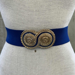 May include: A navy blue elastic belt with a gold-colored, interlocking circular buckle. The buckle has a detailed, rope-like border and a central embossed design. The belt is displayed on a mannequin.