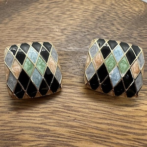 May include: A pair of square earrings with a gold-tone border. Each earring features a black, diamond-patterned design with accents of light blue, green, peach, and gray. The earrings are displayed on a wooden surface.