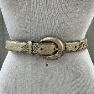 Może przedstawiać: Złoty pasek ze wzorem skóry węża. Pasek ma dużą, zaokrągloną klamrę i jest zapięty wokół manekina. Wzór skóry węża jest w odcieniach beżu i brązu.