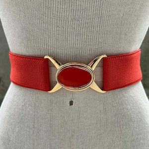 May include: A red elastic belt with a gold-colored buckle. The buckle features an oval-shaped, red-colored center stone. The belt is displayed on a mannequin.