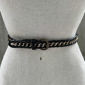 May include: Black belt with a braided leather design and gold chain accents. The belt features a circular, bronze-colored buckle and is displayed on a mannequin. A stylish accessory for any outfit.