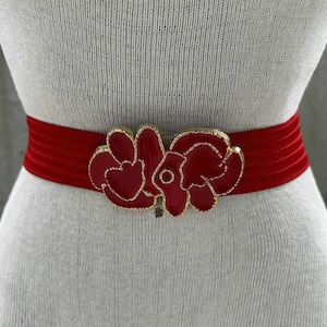 May include: A red velvet belt with a gold-trimmed floral buckle. The buckle features two red flower designs with gold outlines. The belt is displayed on a mannequin.