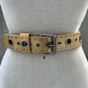 Puede incluir: Cinturón de cuero color beige con múltiples ojales plateados y hebilla plateada. El cinturón se muestra en un maniquí y parece vintage o desgastado. Es un accesorio de moda.