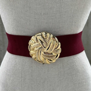May include: A burgundy elastic belt with a large, gold-toned, woven-style buckle. The belt is displayed on a mannequin, showcasing its design and fit. The buckle is the focal point, adding a touch of elegance to the accessory.